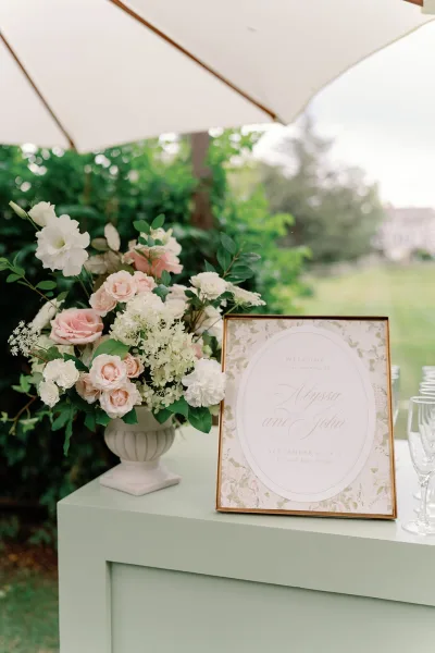 Wedding welcome sign in a gold frame with blush roses, white flowers and greenery in an urn vase on a sage pedestal on the lawn