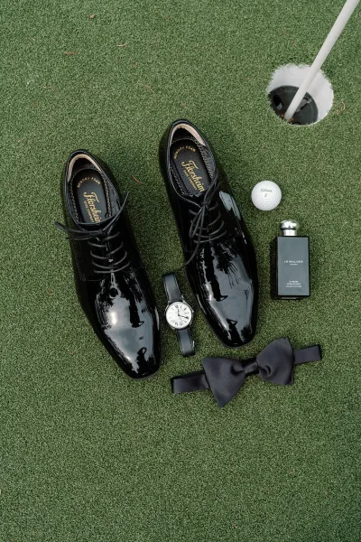 Groom accessories flatlay with black patent shoes, bow tie, watch and cologne beside a golf ball on green turf near a flagstick