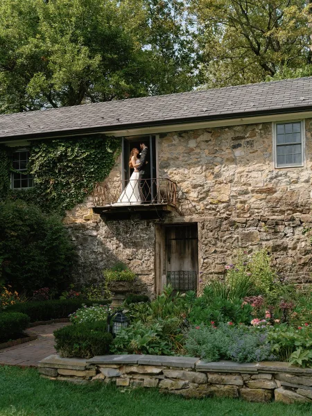 Wedding couple portrait of bride in a strapless gown and groom in a black suit embracing on an ivy-lined balcony by a stone house