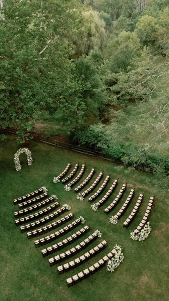 Ceremony setup with curved seating facing a white floral arch on a grassy lawn, greenery aisle arrangements, and trees beyond a fence