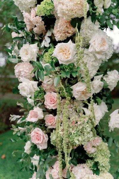 Wedding floral installation with a ceremony flower arch of white and blush roses, hydrangeas, and greenery, cascading on a garden lawn