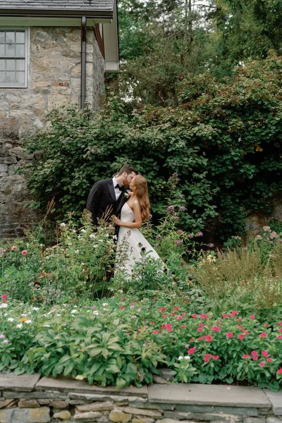 Wedding kiss portrait of bride and groom kissing, her strapless dress and his tuxedo with boutonniere by garden greenery and stone house window