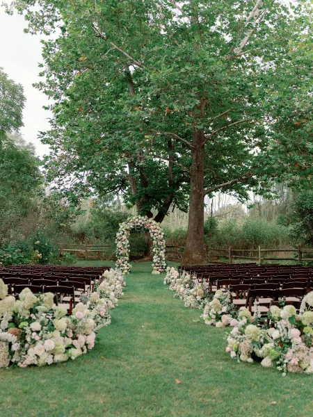 Ceremony setup for an outdoor wedding ceremony with wooden chairs facing a round floral arch on a lawn beneath a large tree