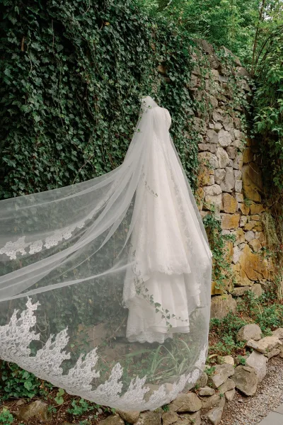 Wedding dress with lace wedding veil hanging outdoors against an ivy-covered stone wall beside a gravel path and greenery