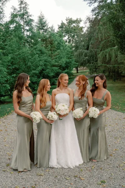 Bride with bridesmaids in sage green dresses holding white rose bouquets, standing on a gravel path with trees behind them