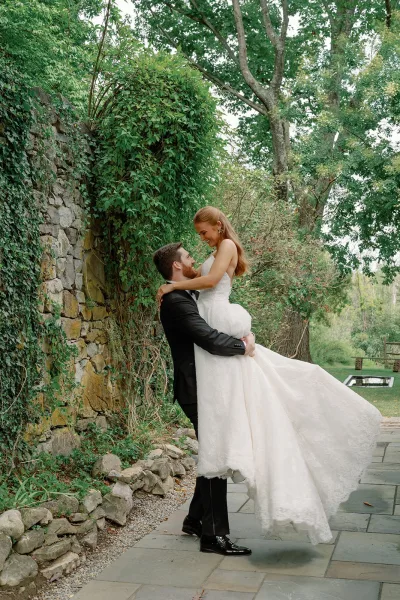 Couple portrait of groom lifting bride in his arms, her veil and dress train flowing beside an ivy-covered stone wall in a garden setting