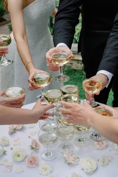 Wedding toast with champagne coupe glasses raised by bride and groom, engagement ring and bracelets visible, greenery and stone steps behind