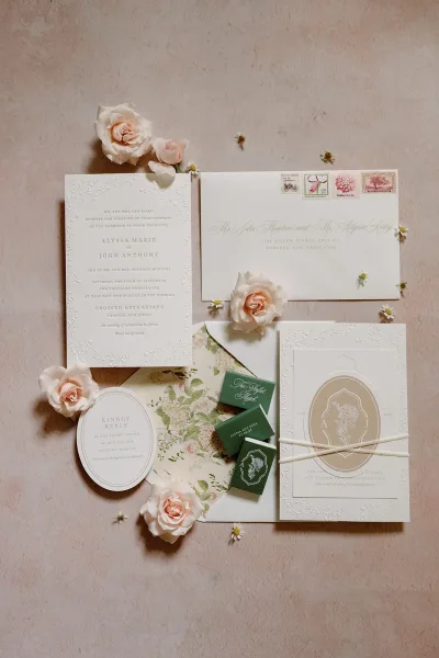 Invitation suite flat lay with embossed wedding invitations, blush roses, floral liner, stamps, wax seal stickers, and ribbon on stone surface