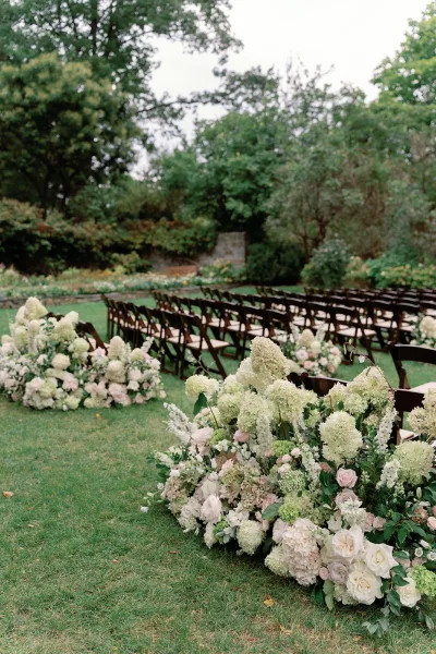 Ceremony aisle decor with hydrangea and rose florals lining a grass path, flanked by wood folding chairs on an overcast garden lawn