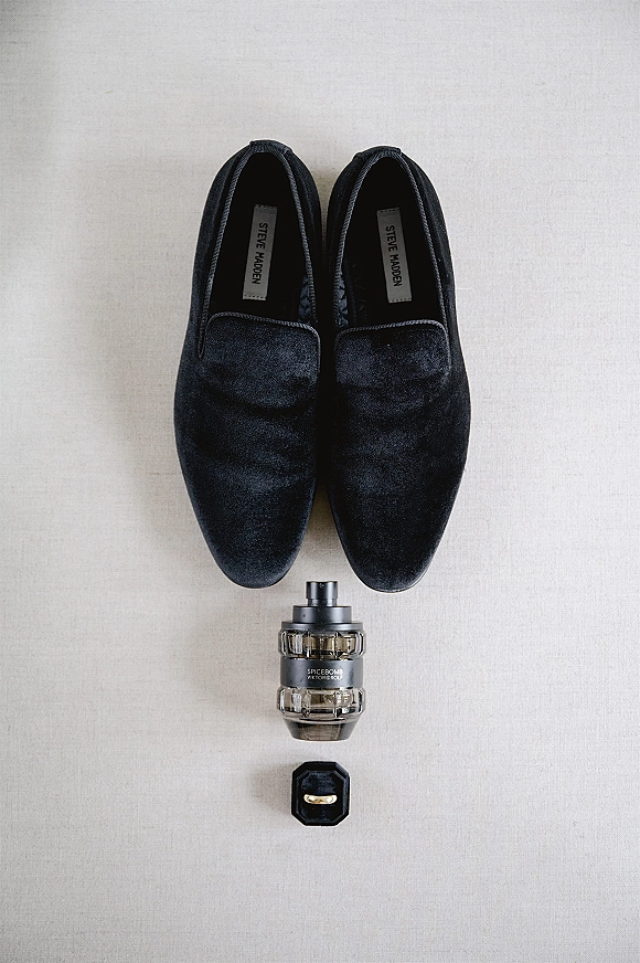 Groom accessories flat lay with velvet loafers, cologne bottle, wedding band, and ring box arranged on a light fabric backdrop