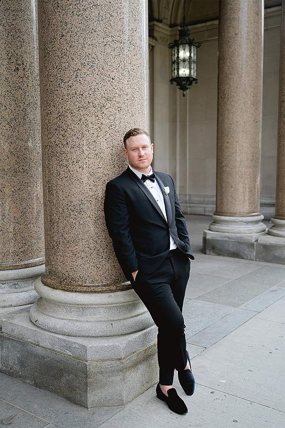 Groom portrait in a black tuxedo look with bow tie and boutonniere, hands in pockets under stone columns and lantern-lit arcade