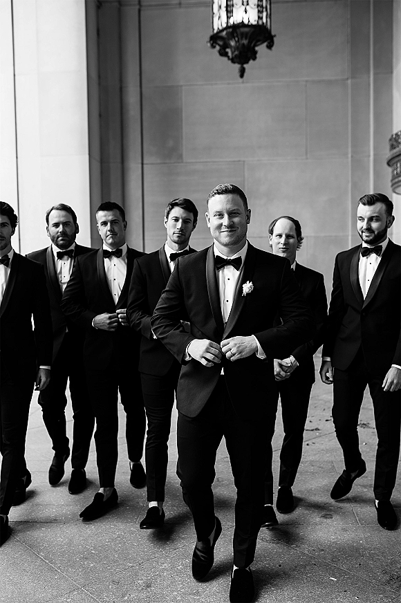 Groom and groomsmen in a groomsmen portrait wearing black tuxedos with bow ties and boutonnieres at a stone-columned entrance with lantern