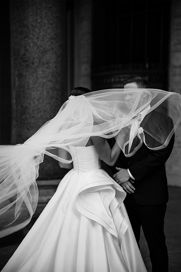 Wedding couple portrait in black and white, bride and groom embrace as her veil lifts in the wind before stone columns at an entrance