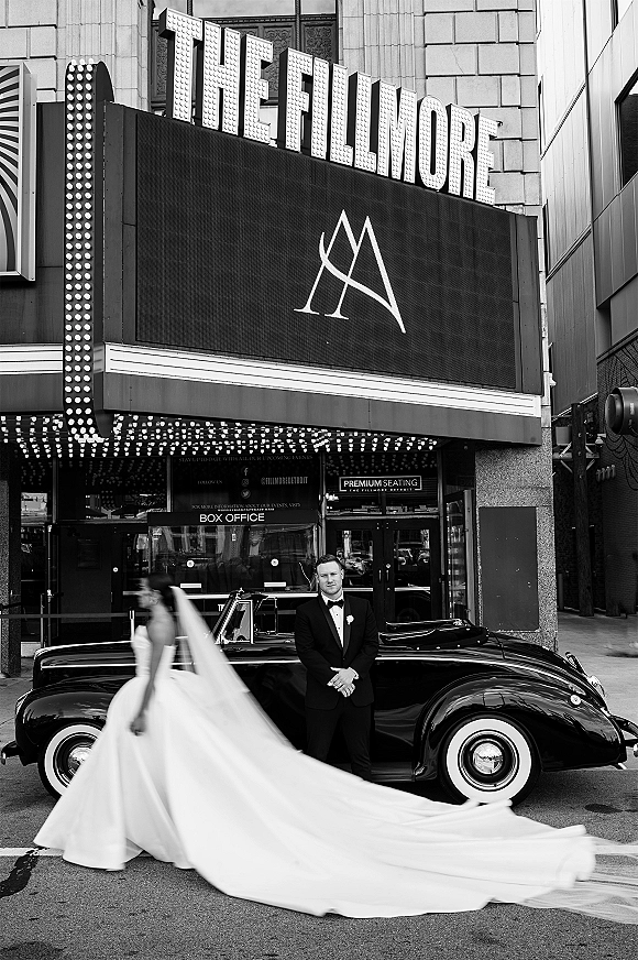 Wedding couple portrait with bride in long train and veil walking past groom in tux by a vintage convertible under theater marquee