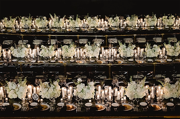 Reception tablescape with white floral centerpieces, pillar candles in glass holders, and gold charger plates on long banquet tables in a dark room