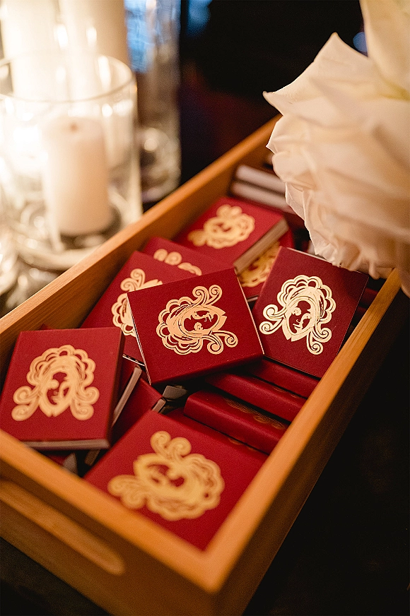 Wedding favors in red and gold favor boxes with a gold monogram, arranged on a wooden tray beside glass pillar candles on a dark tabletop