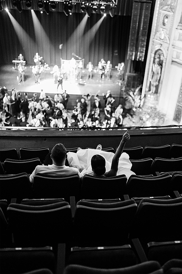 Wedding reception crowd in an ornate theater auditorium with stage lighting, live band on stage, and guests in theater seats watching music