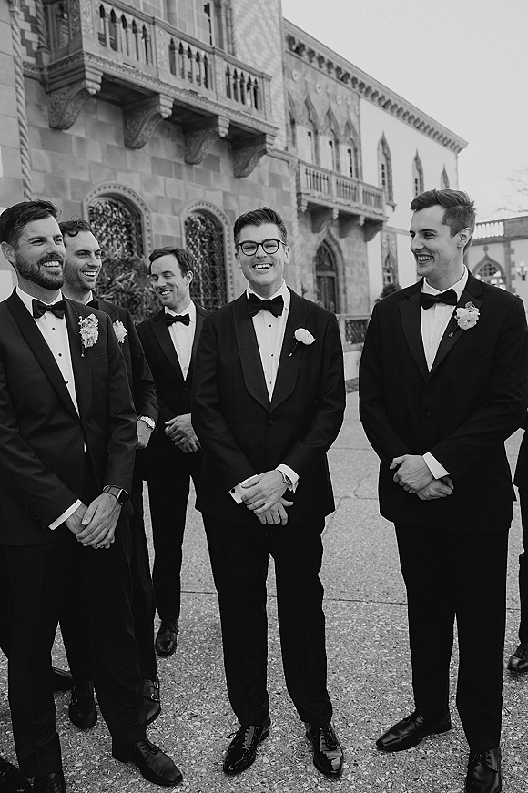 Groomsmen portrait of the groom and groomsmen laughing in black tuxedos with boutonnières in a courtyard beside a stone building with arches