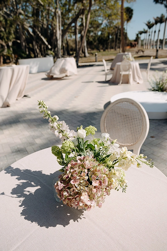 Wedding centerpiece with hydrangea centerpiece in a glass vase on a round table with blush linens, set on an outdoor patio under palms