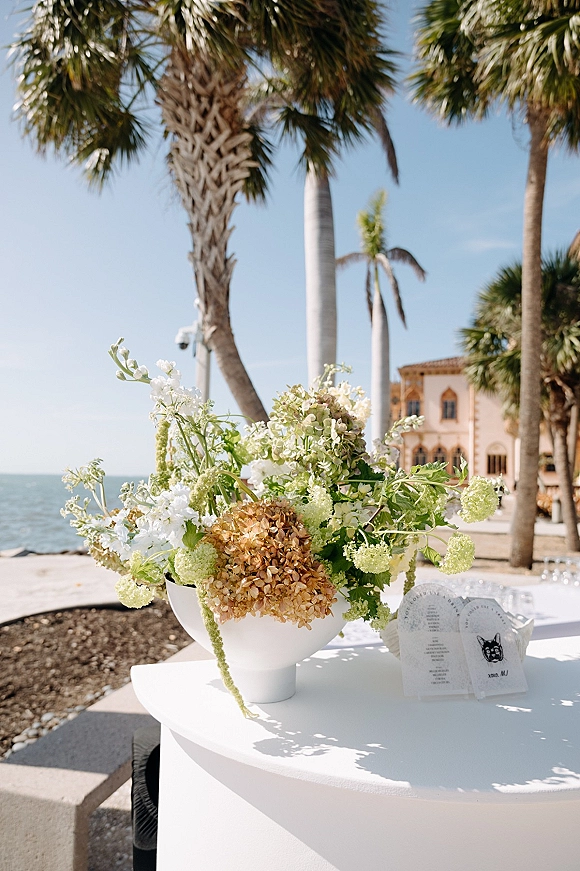 Wedding floral centerpiece in a white pedestal bowl vase beside letterpress escort cards, set against palm trees and ocean on a sandy beach