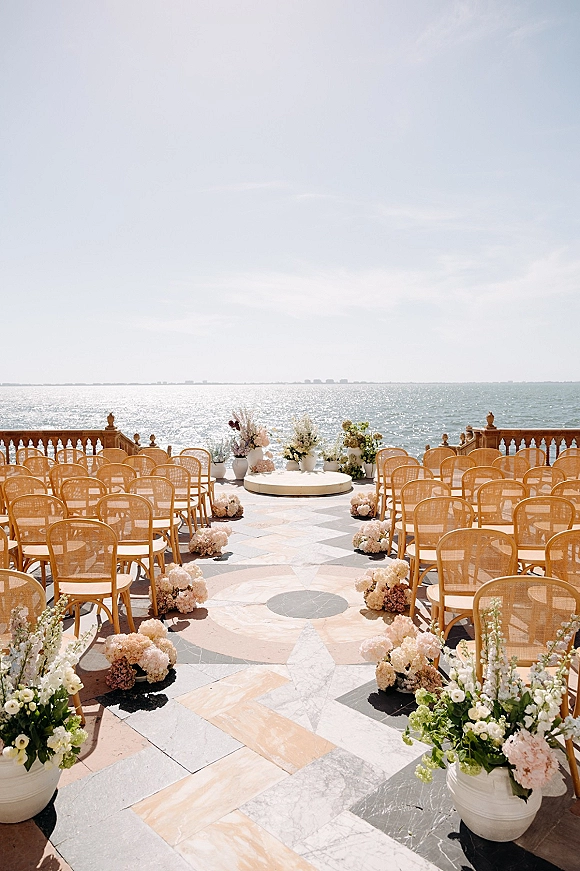 Ceremony setup for an outdoor wedding ceremony with wood chairs and floral aisle clusters on a stone terrace overlooking the ocean view
