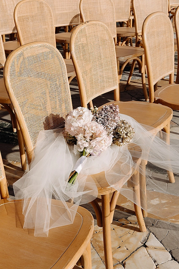 Bridal bouquet of soft hydrangea wedding bouquet blooms with tulle wrap resting on a cane-back chair along stone patio ceremony rows
