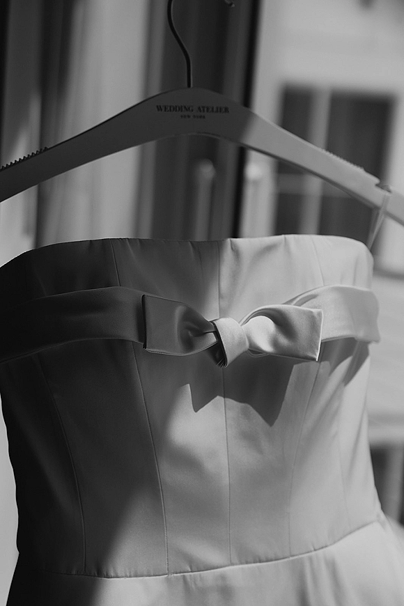 Wedding dress with strapless bodice hanging on a dress hanger, finished with a satin bow, lit by soft window light in a room
