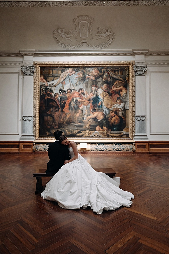 Couple portrait of bride resting on groom shoulder, seated on a bench in an art gallery with a large framed painting backdrop