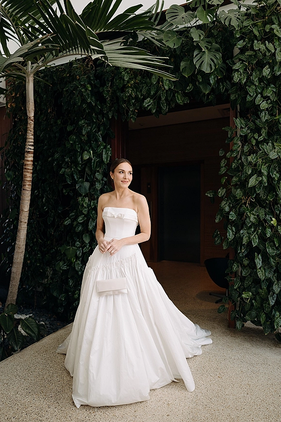 Bridal portrait of a bride in a strapless wedding dress with veil, hands clasped, looking away beside a tropical greenery wall indoors