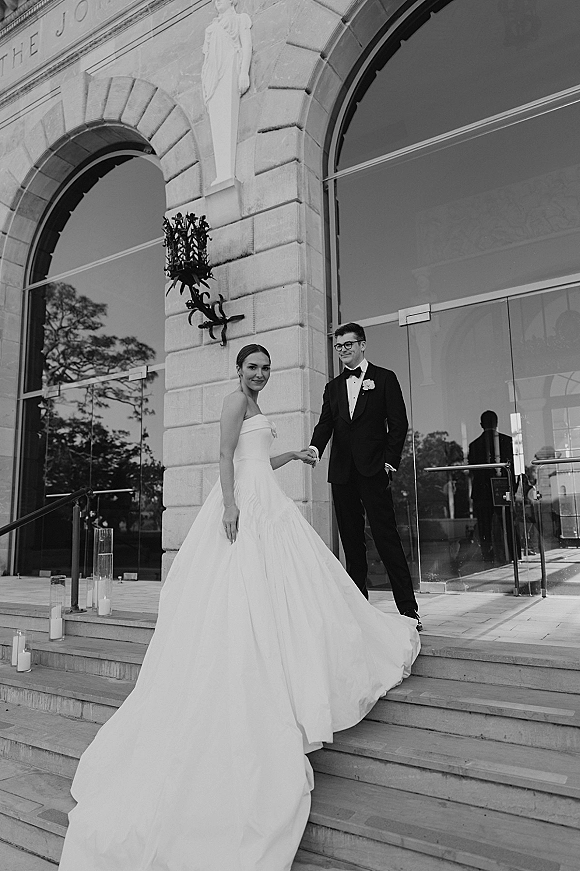 Couple portrait of bride and groom holding hands on stone steps, her strapless gown’s long train flowing beside glass hurricane candles
