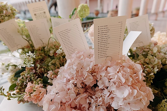 Wedding escort cards and wedding seating chart cards tucked into hydrangeas and greenery on a tabletop display against soft pink walls with railing
