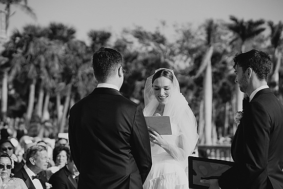 Wedding vows as bride reading vows into a microphone, veil draped over her dress, groom in tuxedo facing her at a palm-lined ceremony