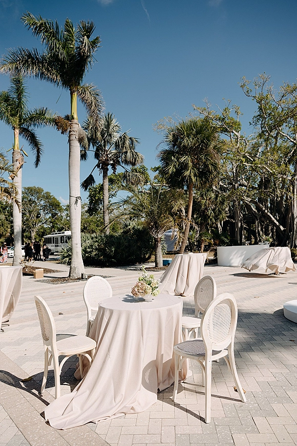 Outdoor reception lounge with round table in draped linens, white bistro chairs and a small floral centerpiece on a palm-lined patio under blue sky