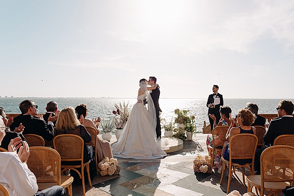 Ceremony kiss as bride in strapless gown and long veil meets groom in black tux beneath floral arch on oceanfront stone terrace
