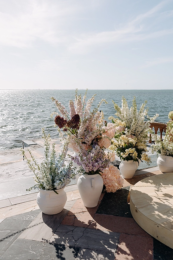 Ceremony florals with aisle floral urns in white ceramic vases, pastel blooms on a wooden stage overlooking the ocean horizon on a stone terrace