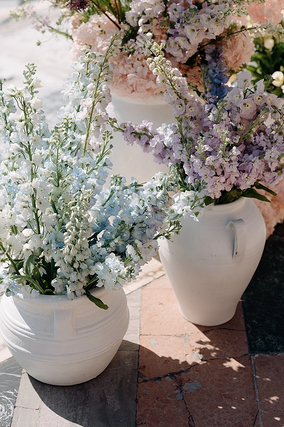 Wedding floral arrangements in white ceramic vases with pastel delphinium, hydrangea, and greenery on a sunlit stone patio with shadows