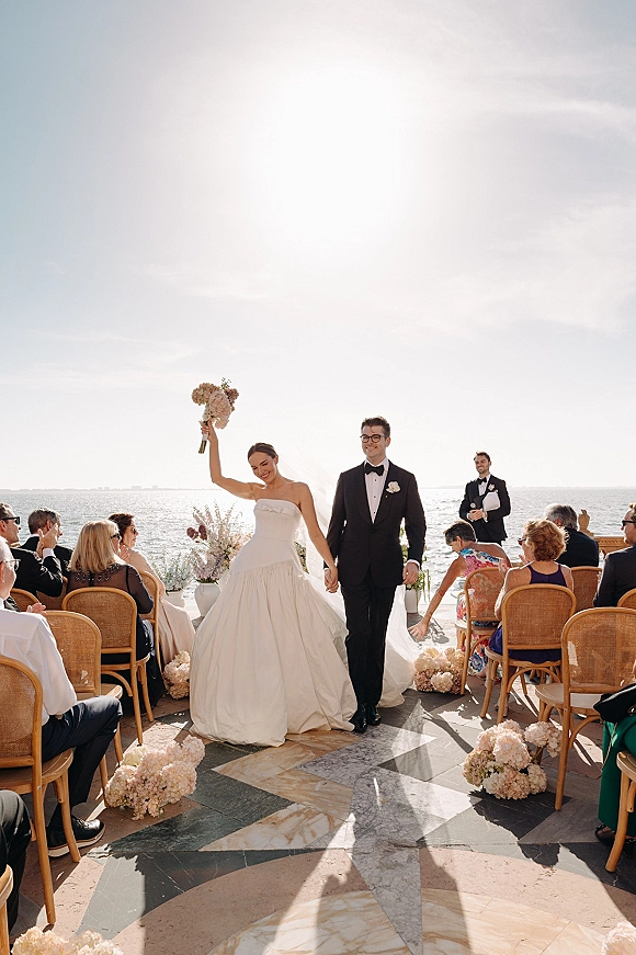 Wedding recessional with bride and groom walking aisle, holding hands as she lifts bouquet on a sunlit oceanfront stone patio.