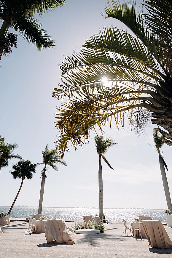Outdoor reception setup with waterfront wedding reception round tables in beige linens, white chairs, and low floral centerpieces by palm trees and ocean