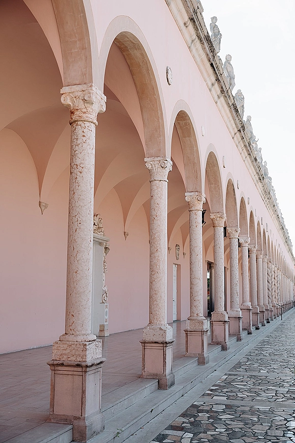 Wedding venue architecture with arched colonnade wedding venue, stone columns and statues along a pink covered walkway with mosaic pavement