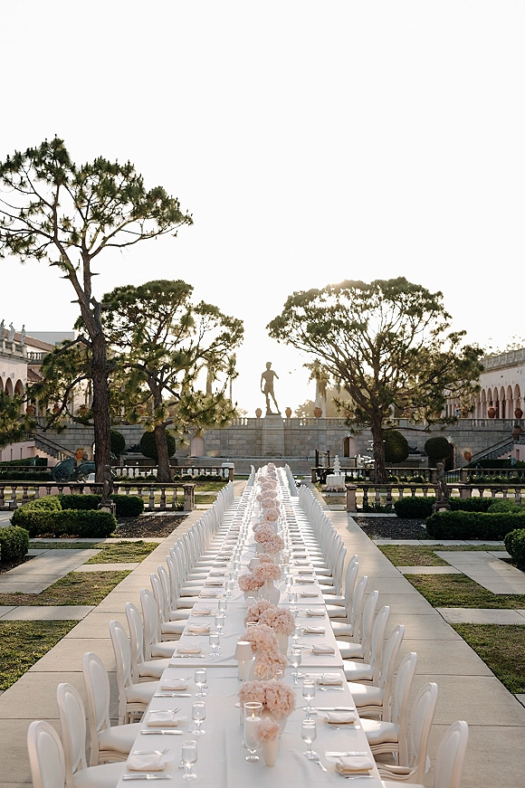 Reception tablescape with long wedding banquet table in a formal garden courtyard, blush floral centerpieces, candles, and white chairs on linen