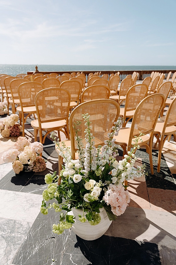 Ceremony setup with rattan bistro chairs and aisle florals, featuring white urn hydrangeas and roses on an oceanfront terrace deck