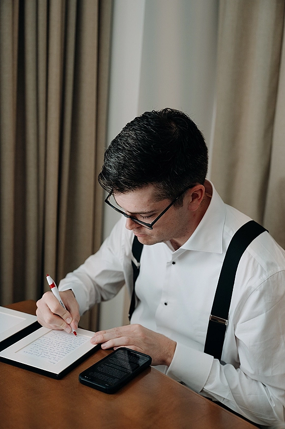 Groom getting ready, writing vows in a notebook at a wooden table, wearing a white dress shirt and black suspenders by window light