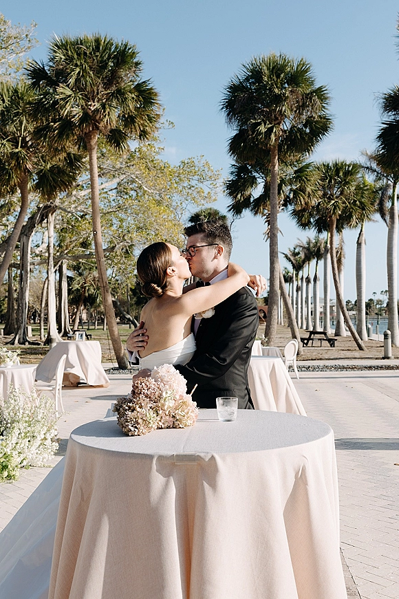 Wedding kiss as bride in strapless gown and groom in black tuxedo embrace on a waterfront patio with palm trees and blue sky backdrop