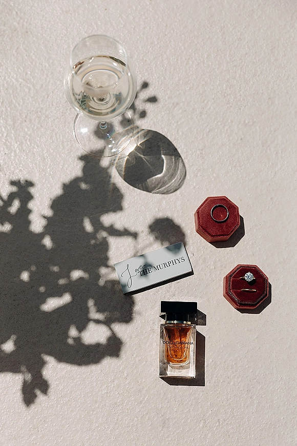 Wedding rings flatlay with engagement ring and band beside a place card and perfume bottle on a white textured surface with floral shadows