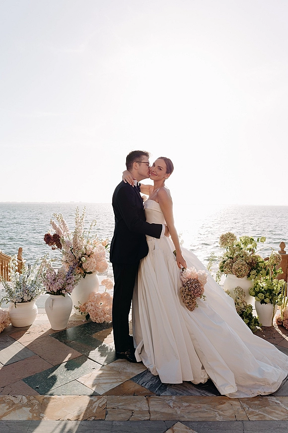 Couple portrait of bride and groom embrace, groom kissing her cheek as she looks at camera on an oceanfront terrace, bouquet in hand