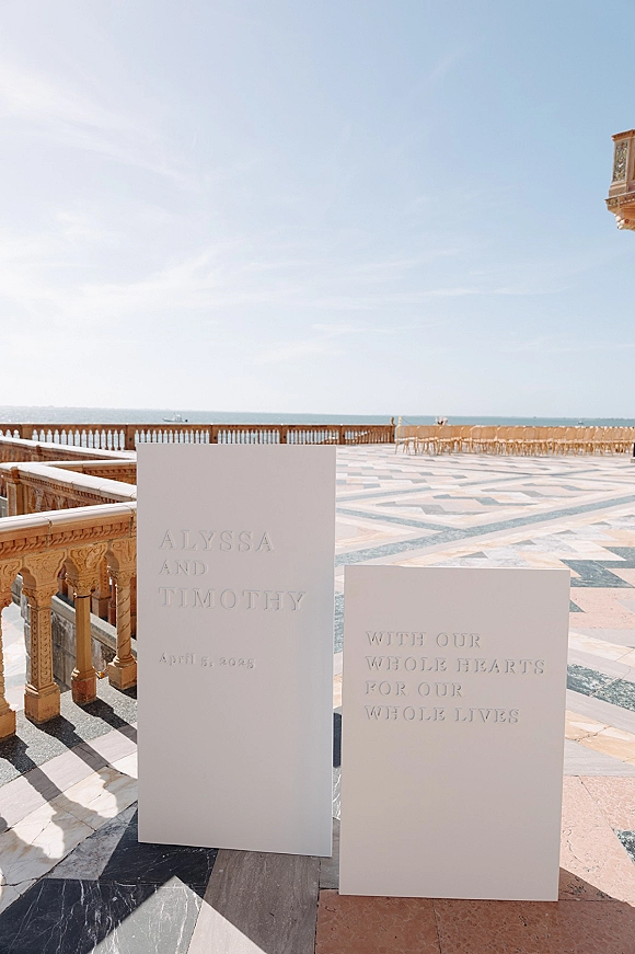 Wedding welcome sign with embossed wedding sign details on freestanding white boards, set on a stone terrace overlooking the ocean horizon