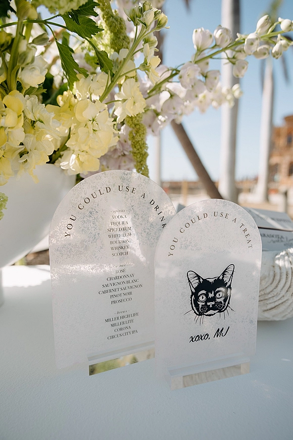 Wedding bar menu acrylic bar menu sign on wooden stands beside white flowers and greenery, set outdoors under blue sky with columns