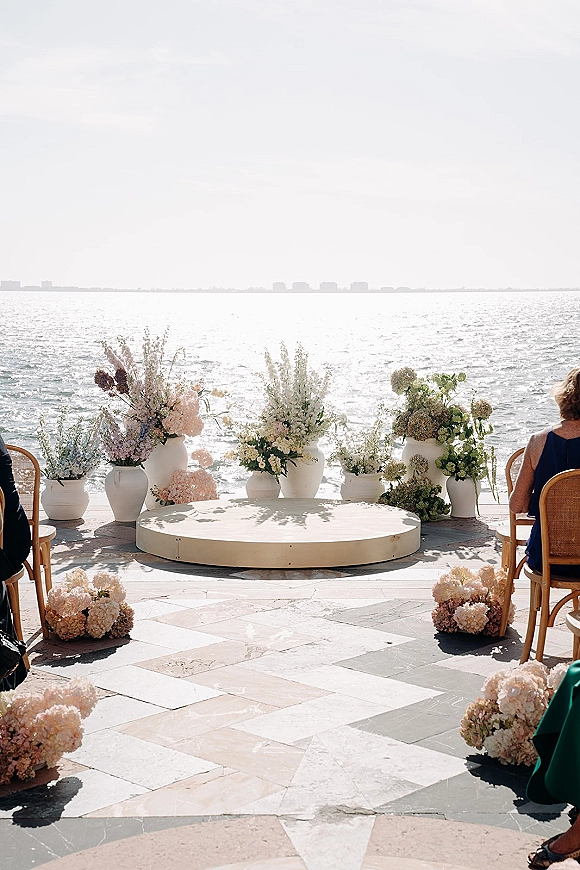 Ceremony setup with oceanfront wedding ceremony flowers in white ceramic vases, round platform, and wooden bistro chairs on a stone patio.