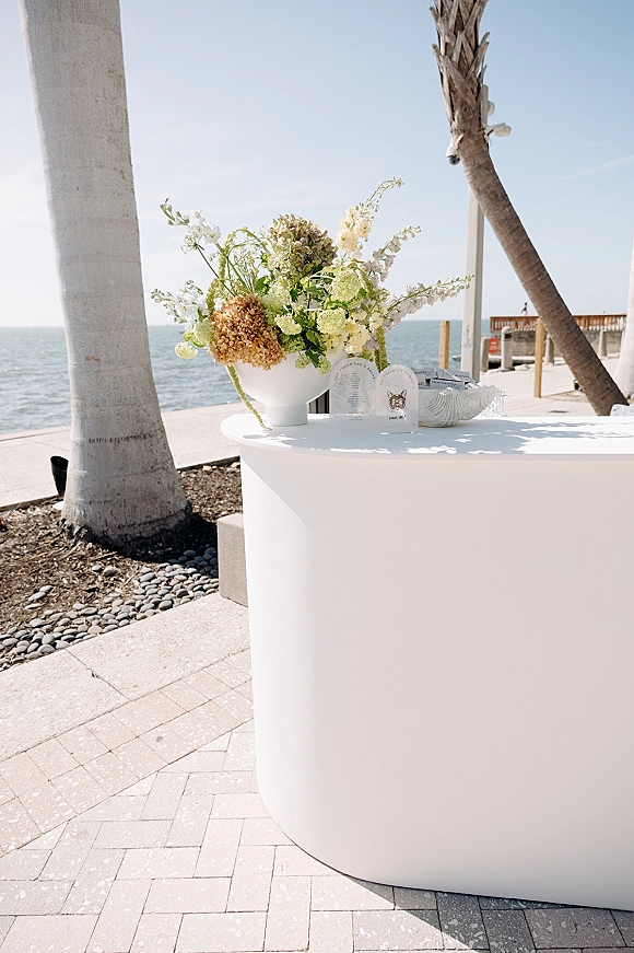 Escort card display with wedding escort cards beside a white bowl vase floral arrangement on pedestal table, with ocean and palm trees behind