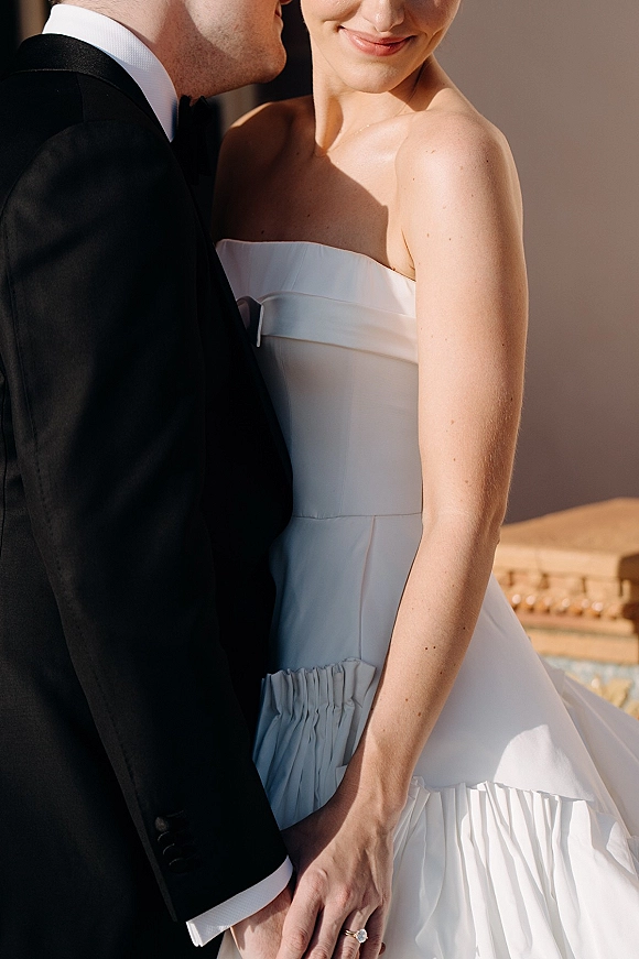 Wedding couple portrait of groom in black tuxedo kissing bride’s cheek, her strapless dress and engagement ring glowing in outdoor light